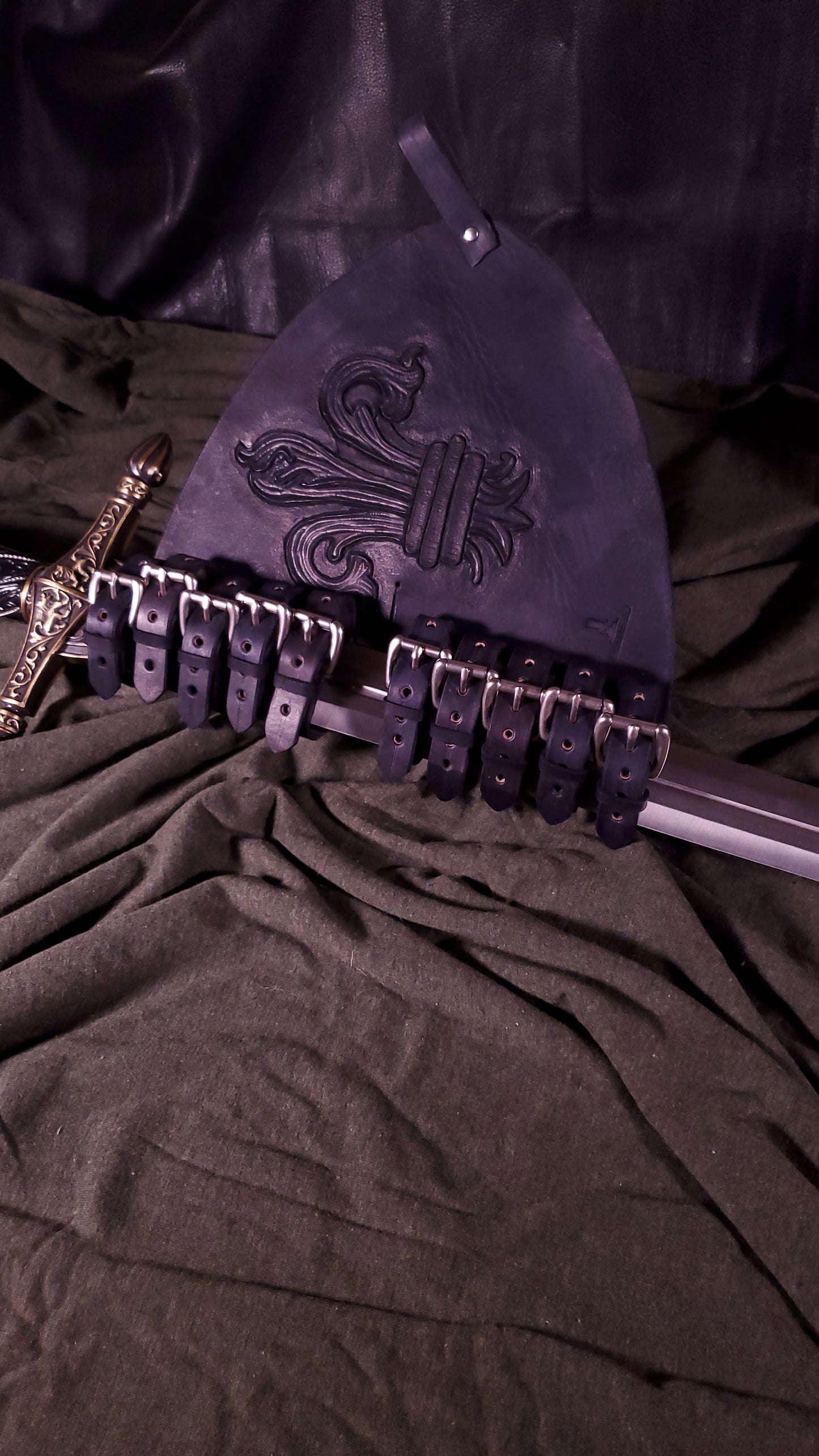 A sword carrier with a longsword in it. the sword carrier has a large Fleur De Lis tooled on the face of it. It also has two belt loops at the top and side. It also has ten nickel buckles where the sword goes into to adjust for your specific sword.